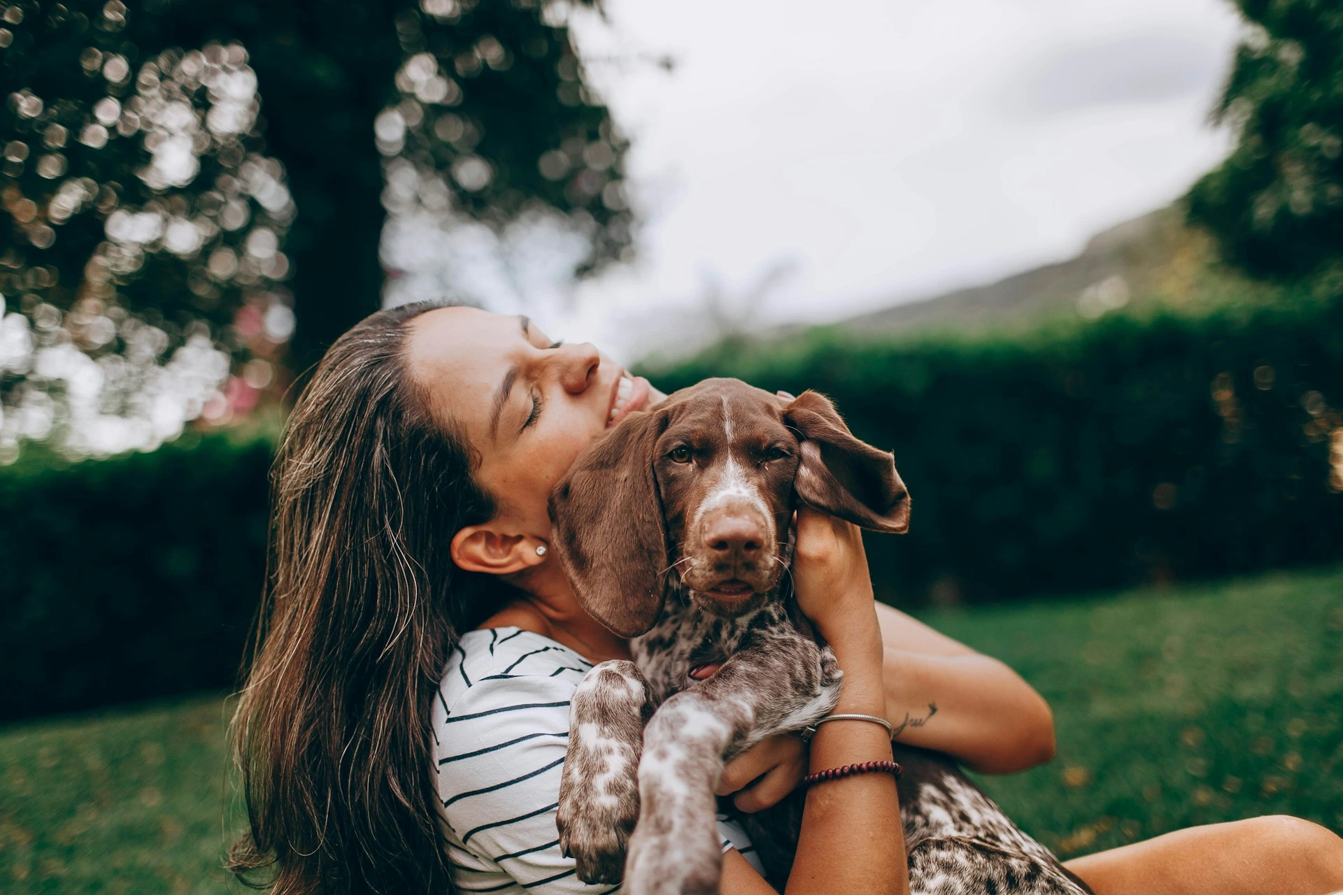 A woman hugs a German Shorthaired Pointer puppy while sitting in a grassy area.